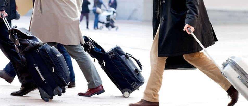 Attention all passengers: In Terminal D, we observe a quartet of travelers advancing in unison, their rolling suitcases obediently trailing behind them. The photographic frame zeroes in on their legs and the luggage wheels smoothly navigating the paved concourse. Meanwhile, a gently blurred backdrop of bustling voyagers including a stroller-bound child adds depth to our scene. Please ensure your belongings are with you at all times for your journey through Dallas/Fort Worth International Airport.