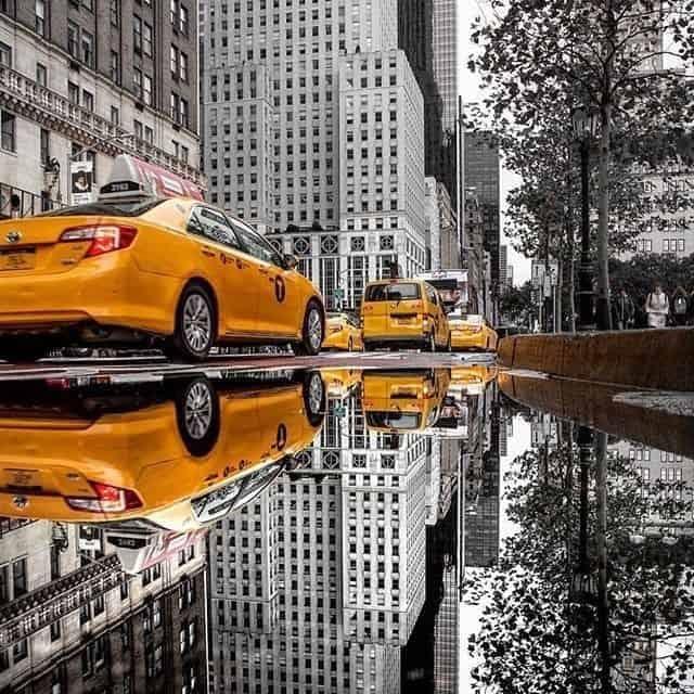 Yellow taxis, including an Eagle Mountain taxi cab, reflected in a puddle on a busy city street with tall buildings and trees in the background.