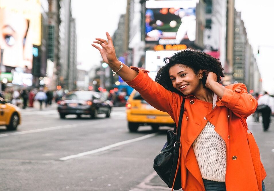 Scene: A bustling cityscape, the air punctuated by the hum of urban life. Amidst the orchestrated chaos, a woman adorns an orange coat of radiant hue, curls cascading down her shoulders. Her smile beams as she confidently raises a hand to secure her DFW airport transfer. The street teems with movement. Yellow cabs crisscross in vivid streams, while pedestrians navigate their paths with rhythmic precision. Above this choreography of motion, electronic billboards ignite the scene with vibrant illuminations, casting their digital glow across the urban canvas.